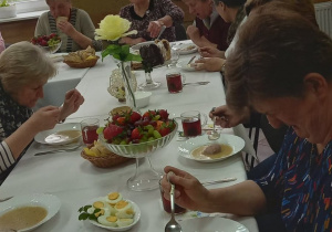 Seniorzy z Klubu Senior+ siedzą przy stole wielkanocnym i jedzą żurek z białą kiełbasą i jajkiem.