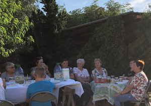 Seniorzy siedzą przy stole i jedzą kiełbaski z ogniska.
