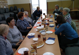 Seniorzy będący uczestnikami Klubu "Senior+" oraz Pani Kierownik Gminnego Ośrodka Pomocy Społecznej siedzą przy stole podczas poczęstunku z okazji Międzynarodowego Dnia Osób Starszych.