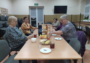 Seniorzy będący uczestnikami Klubu "Senior+" siedzą przy stole podczas poczęstunku z okazji Międzynarodowego Dnia Osób Starszych.