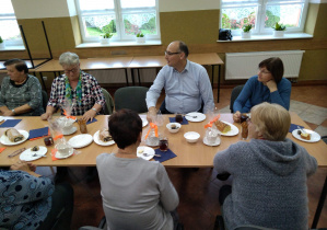 Seniorzy będący uczestnikami Klubu "Senior+", Pan Wójt oraz Pani Kierownik Gminnego Ośrodka Pomocy Społecznej siedzą przy stole podczas poczęstunku z okazji Międzynarodowego Dnia Osób Starszych.