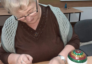 Seniorka będąca uczestnikiem Klubu "Senior+" siedzi przy stole podczas wykonywania ozdoby świątecznej.