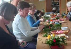 Seniorzy z Klubu Senior+ siedzą przy stole i przygotowują ozdoby wielkanocne.
